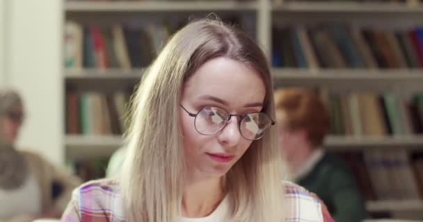 在镜头前 年轻的金发白人女学生戴着眼镜 看着镜头 面带微笑 非常快乐 在图书馆学习的漂亮女人 肖像画 — 图库视频影像