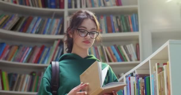 年轻美丽的白种人女学生戴着眼镜 背着背包站在图书馆里 阅读课本并把它关上 把书放在架子上然后离开 文学概念 靠近点 — 图库视频影像