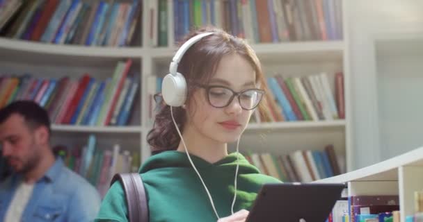 在图书馆 用平板设备和按键在屏幕上关闭眼镜和耳机中的年轻女学生 时尚女性在电脑上发短信 在网上冲浪以获取信息 — 图库视频影像