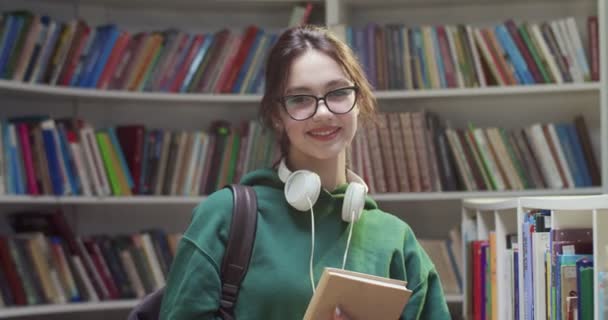 照片中年轻的白种人漂亮的女学生 戴着眼镜 对着相机微笑 并在图书馆里拿着课本 手里拿着书的女人站在书架上 文学概念 — 图库视频影像