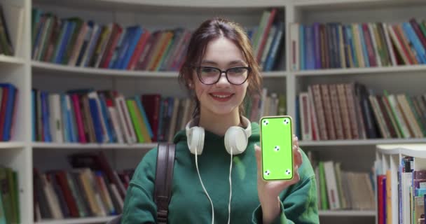 图片说明了年轻的白种人漂亮的女学生戴着眼镜微笑 在图书馆里展示带有绿色屏幕的智能手机和跟踪动作 拿着手机示威的妇女 — 图库视频影像