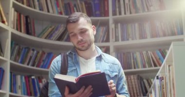 Sırt çantalı yakışıklı beyaz erkek öğrenci kütüphanede duruyor, ders kitabı okuyor ve kapatıyor. Kitabı rafa kaldırıyorum ve gidiyorum. Edebiyat konsepti. Rafların arkaplanı.