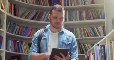 Kafkasyalı genç erkek öğrenci kütüphanede duruyor, tablet cihazı kullanıyor ve ekrana vuruyor. İnternette bilgi almak için internette sörf yapan yakışıklı bir adam..