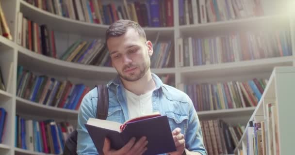 年轻英俊的白种人男生背着背包站在图书馆里 阅读课本并把它关上 把书放在架子上然后离开 文学概念 书架背景 — 图库视频影像