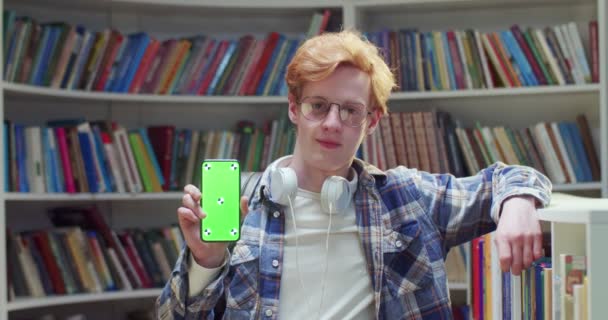在图书馆里 年轻的白种人红头发男生戴着眼镜 笑着展示带有绿色屏幕的智能手机和跟踪动作的肖像 拿着手机示威的家伙 — 图库视频影像