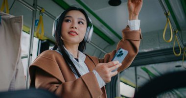 Tren vagonunun veya otobüsün ortasında duran güzel Çinli kız. Bir elinle dengeyi sağlarken diğer elinle akıllı telefonu kullanıyorsun. Kablosuz kulaklıkla müzik dinlemek. Telefon ekranına bağlanılıyor.