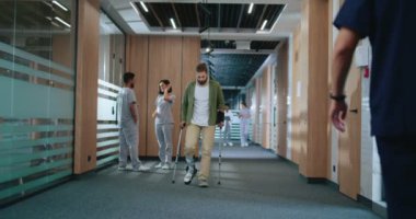 Yaralı beyaz erkek, hastanede düşerken koltuk değneği kullanıyor. Yardım ve destek için koşan güzel genç doktorlar ve hemşireler. Modern hastane kavramı.