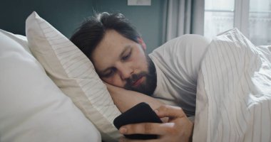 Üzgün, çekici genç adam yatakta uzanıyor, cep telefonuyla mesajlaşıyor. Akıllı telefonla mesaj yaz, iletişim kurmak için uygulamaları kullan.