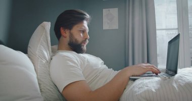 Ciddi bir beyaz adam yatakta yatıyor ve çalışmak için dizüstü bilgisayar kullanıyor. Genç erkek yatak odasındaki bilgisayar klavyesinde daktilo kullanıyor..