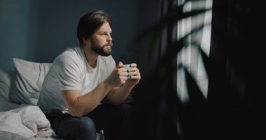 Endişeli Kafkasyalı bir adam bir fincan çayla pencerenin yanındaki yatakta oturuyor. Genç erkek sıcak kahve içiyor ve manzaraya bakıyor..