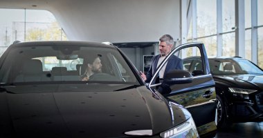 Yeni alınmış bir arabanın içinde oturan Kafkasyalı alıcı otomobil kuaförüne bakıyor. Tabletli bir satıcı, ilgili alıcıya lüks bir arabadan bahsediyor..