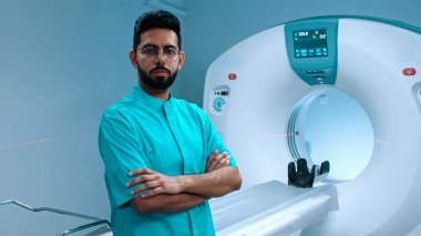 Hintli tomografi uzmanı çapraz kollarla poz veriyor. CT tarayıcısının arka planında kendinden emin bir doktor var. Doktorun gözlük taktığı boy. MRI incelemesi için modern donanımlı oda. MRI kapsülü.