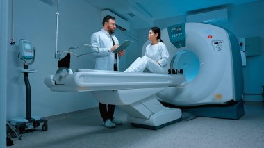 MRI kapsülü ve klinikte yeni modern ekipmanlar. Modern klinikte biyopsi için röntgen bekleyen Asyalı kadın hasta. Tarayıcı masasında otururken doktoruna mide ağrısı şikayeti..