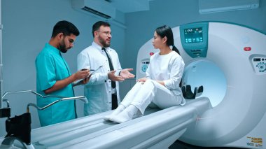 Profesyonel doktor ve üniformalı hemşire hastanın yanında duruyor ve prosedürü açıklıyor. MRI kapsülü ve klinikte yeni modern ekipmanlar. Tıbbi personel konuşurken hasta dikkatlice dinliyor..