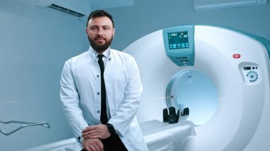 Kendine güvenen doktor manyetik rezonans görüntüleme kapsülünde oturuyor. Gözlüklü sakallı doktor. MRI taraması için modern donanımlı oda. MRI kapsülünün arka planında beyaz önlüklü doktor var..