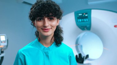 Tomografi uzmanının geniş gülüşlü yakın çekimi. MRI kapsülünün arka planında gülümseyen neşeli doktor. MRI incelemesi için kabine. Tomografi teknisyeni. Manyetik rezonans görüntüleme.