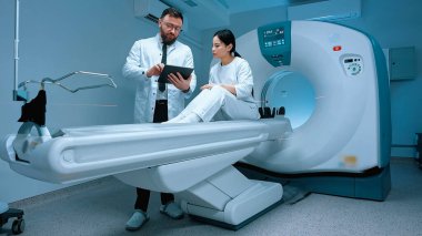 MR çekilmeden önce doktor hastayla konuşuyor. Doktor tableti tutuyor. Hasta tomografide oturuyor. Doktor MRI işleminden önce talimat veriyor. Manyetik rezonans görüntüleme. Doktor ve hasta..