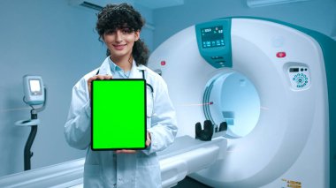 Genç tomografi uzmanı elinde krom anahtarlı tablet tutuyor. MRI cihazlarının arka planında gülümseyen neşeli bir doktor. MRI incelemesi için kabine. Yeşil şablonlu tablet gösteren bir tıp çalışanı.