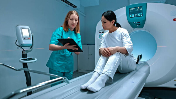 Doctor holding tablet and showing result of MRI. Patient lying at MRI capsule. MRI cabinet. Modern equipment in private clinic. Doctor talking to patient after procedure. MRI examination.