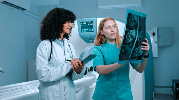 Chef doctor holding MRI scan. Intern with stethoscope holding folder and making notes. Two tomographers in cabinet of MRI scanning. Capsule of MRI examination. Machine for CT scanning.