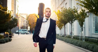 Beyaz erkek portresi arkasında gitarla takım elbise giyiyor. Yakışıklı adam sabah güneşiyle sokak lambasında yürür..