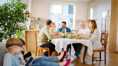 Mutlu aile yemek odasında öğle yemeği yiyerek harika zaman geçiriyor. Akrabalar lezzetli yiyeceklerle dolu masada otururlar. Çocuklar telefondaki uygulamaları kullanarak birbirleriyle oynuyorlar..