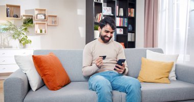 Tatmin olmuş Arap erkek elinde banka kartıyla koltukta oturuyor. Sakallı adam online ödemenin başarılı olmasına sevindi. Erkek doğrudan evden online alışveriş yapmış..