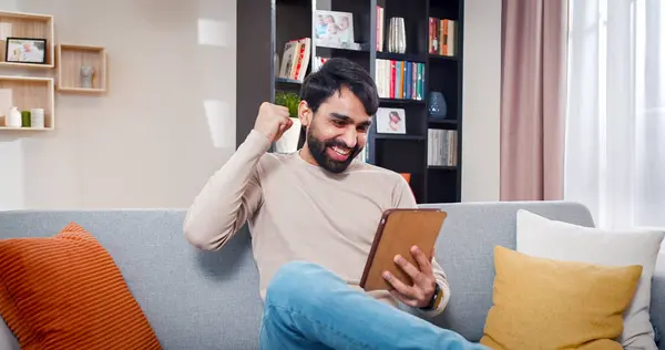 stock image Portrait of Arabian male sitting on sofa looks at tablet screen. Happy man won playing new online game at home. Male is filled with joyful emotions.