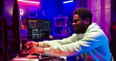 Boynunda kulaklık olan Afrikalı sakallı adamın portresi LED ışıkla aydınlatılmış bir odada bilgisayar ekranının önünde oturuyor. Yakışıklı adam evde DJ ekipmanı kullanıyor..