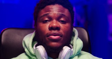 Boynunda kulaklık olan sakallı Afrikalı erkek portresi. Yakışıklı adam LED ışığıyla aydınlatılmış bir odada oturuyor..