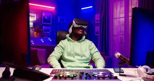 stock image Portrait of African male wearing VR-glasses in room illuminated by LED light. Handsome bearded face man sits at table with different gadgets.