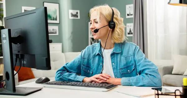 stock image Close up portrait of beautiful Caucasian female wearing headphones with microphone sits at table in front of computer screen. Happy young woman works conducting online meeting at home.