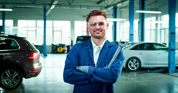 stock image Close up portrait of young Caucasian male wearing protective glasses with working form stands with folded hands smiling. Handsome man stands in the middle of service station with shifting spanner.