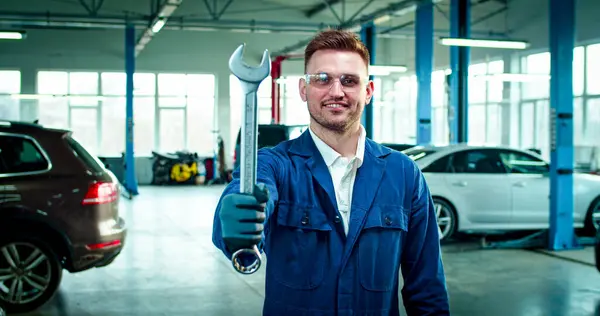 stock image Close up portrait of Young Caucasian male wearing protective glasses with working form stands in the middle of service station with shifting spanner in hand. Handsome man smiles at camera.