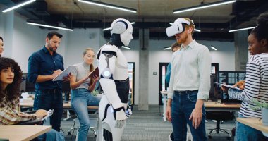 Yeni eserlerini çevreleyen çok ırklı mühendisler ve programcılar. VR kulaklıklı bir erkek hareket ediyor ve insansı robotu çalıştırıyor. Sinerji kontrol ediliyor. Deneme. İnsanlar başarıdan mutlu..
