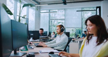 Beyaz erkek mikrofonlu kulaklığı alıyor. Müşteriyle konuşuyor. İnsanları desteklemek. Bilgi ya da yardım sağlıyorum. Merkezi ara. Danışmanlık ya da mal satma. Çalışırken neşeyle gülümsüyor.