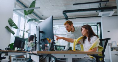 Attractive man helping his coworker solving work issues. Pretty woman sitting at desk with computer while manager standing behind. Female explaining to man something and pointing finger on screen. clipart