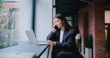Büyük ofis binasının koridorunda büyük pencerenin önünde oturan güzel Asyalı kız. Tek elle klavyede yazarken akıllı telefondan konuşmak. Kadın iş toplantısından önce konsantre oldu.