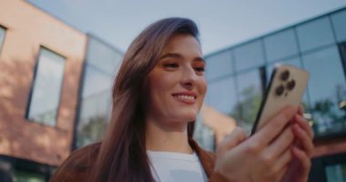 Dışarıda yürüyen ve iki eliyle mobil cihaz kullanan hoş beyaz bir kadın. Mesaja cevap verirken kendi kendine gülümsüyor. Metni dokunmatik ekrandan giriyorum. Akıllı telefona odaklanarak bakıyorum..