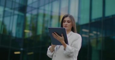 Güzel beyaz kadın ofis binasının önünde duruyor ve tablet cihaz kullanıyor. Taşınabilir bilgisayarın ekranında yazıyorum. Arkadaşlara ya da iş arkadaşlarına mesaj atmak ya da mesajlaşmak. Müdür molada çalışıyor.