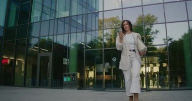 Beyaz kadının büyük iş merkezinden çıkarken ön kamera görüntüsü. Bir elinde büyük, şık bir çanta tutuyor. Akıllı telefonun dokunmatik ekranında yazıyorum. İşten sonra evden ayrılmak..