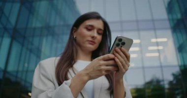Kamera, beyaz kadın tarzında hareket ediyor. İki eliyle akıllı telefon kullanan bir kadın büyük ofis binasının altında duruyor. Uzaktan iletişim konsepti. Modern teknoloji. Web taraması.