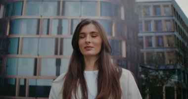 Çekici beyaz kadın ortada duruyor ve doğrudan kameraya bakıyor. Poz verirken gülümsüyor. Modern mimari bulanık odak noktasında kadının arkasında görünür. Şık kıyafetler..