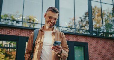 Güzel görünümlü beyaz adam meydanda yürürken etrafına bakıyor. Bir yere giderken akıllı telefondan mesaj yazan tatmin olmuş bir erkek. Teknoloji ve gülümseme kullanarak..