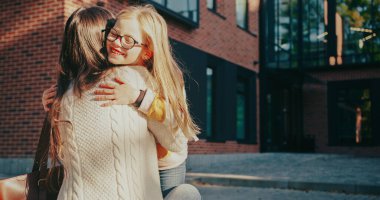 Kameranın yakın görüntüsünde sarı saçlı ve gözlüklü beyaz bir kız annesine sarılıyor. Sıcak bir andan beri gülümsüyor. Çocukların yüzünde güneş parlıyor. Okulu bitirdiğim için mutluyum..