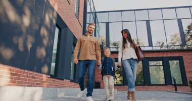 Modern mimariyle tuğla binanın önünde yürüyen mutlu bir aile. Oğlunun elini tutan uzun saçlı güzel bir anne. Diğer taraftaki baba neşeli görünüyor. Ortadaki çocuk sırt çantası taşıyor.