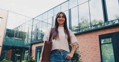 Modern ticaret bürosunun önünde büyük pencereli, en iyi manzara için poz veren şık beyaz kadın. Kameraya bakıp gülümsüyorsun. Bir elini kot pantolonun cebinde tutmak. İşten sonra okuldan ayrılmak..