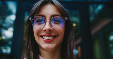 Kafkasyalı, yuvarlak gözlüklü bir kadının yan taraftaki bir şeye baktığı çok yakından görülüyor. Doğrudan kameraya bakıyorum. Yüzünde sıcak bir gülümseme var. Arka plandaki uzun pencerelerde görünen ağaçlar.