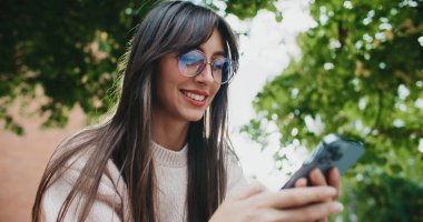 Gözlüklü, beyaz, çekici bir kız temiz havada vakit geçiriyor. Akıllı telefondan arkadaşlarla mesajlaşmak. Uzaktan iletişim için cep telefonu kullanılıyor. Olumlu mesajdan gülümsüyor. Ekranda yazılıyor.