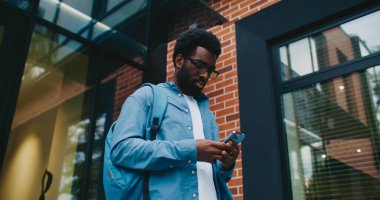 Kamera, sırt çantalı ve mavi gömlekli Afrikalı Amerikalı öğrenciye yaklaşıyor. Tuğla binanın yanında duran ve iki eliyle akıllı telefon kullanan bir adam. Ekranda iki parmakla yazmak.